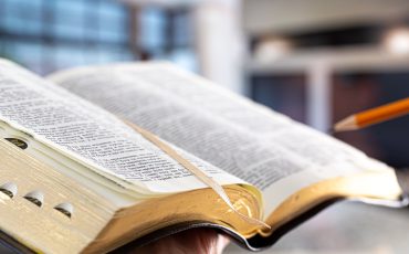 A man holds a Bible with a pencil, against the background of the living room.